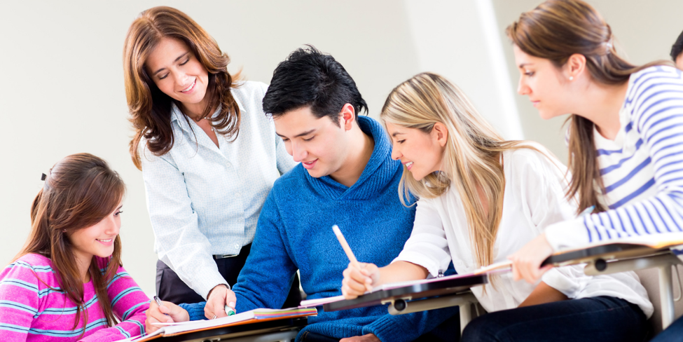 Group of 3 female students and 1 male student smiling while teacher points at work in male student's binder.